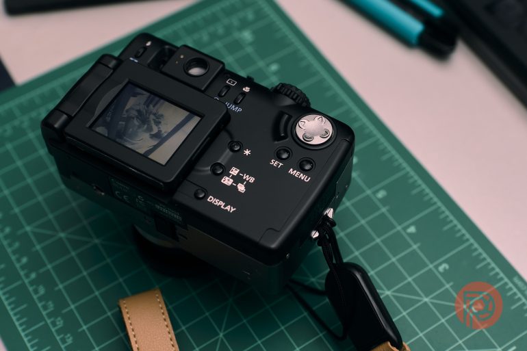 Black digital camera on a green cutting mat, displaying a photo on its screen, strap attached.