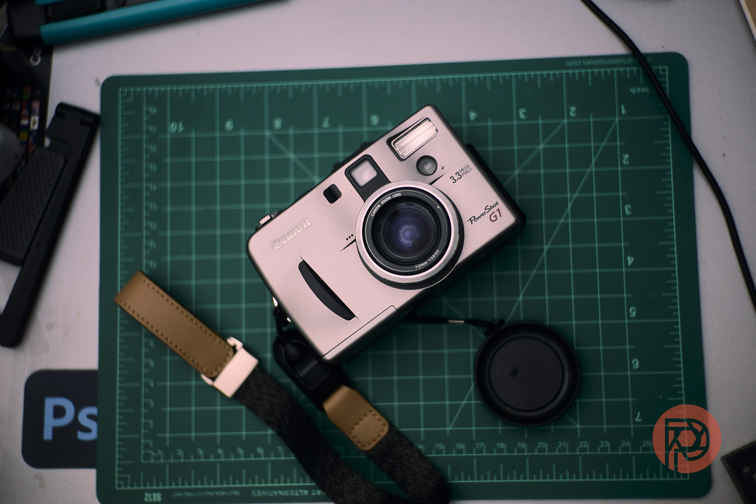 Silver digital camera on a green cutting mat, with a lens cap and a brown strap beside it.