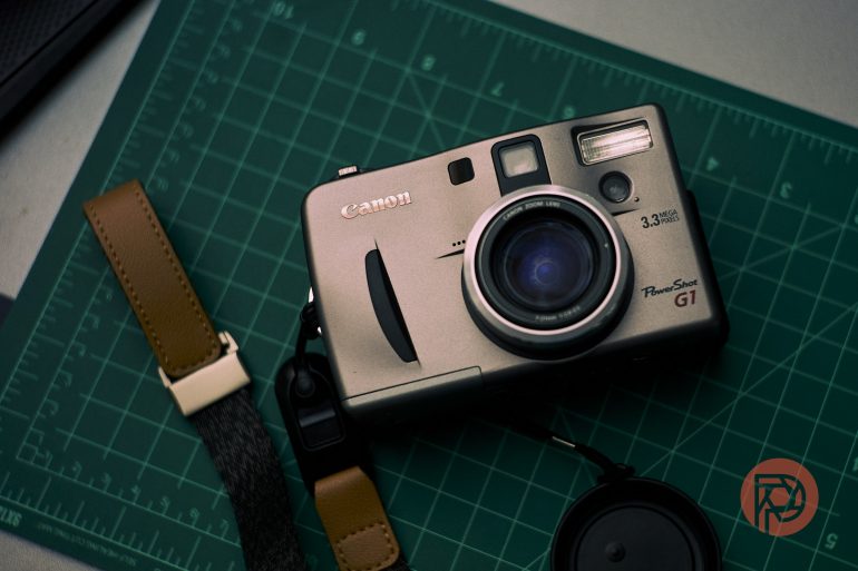 A Canon PowerShot G1 camera on a green cutting mat with a leather strap and lens cap nearby.