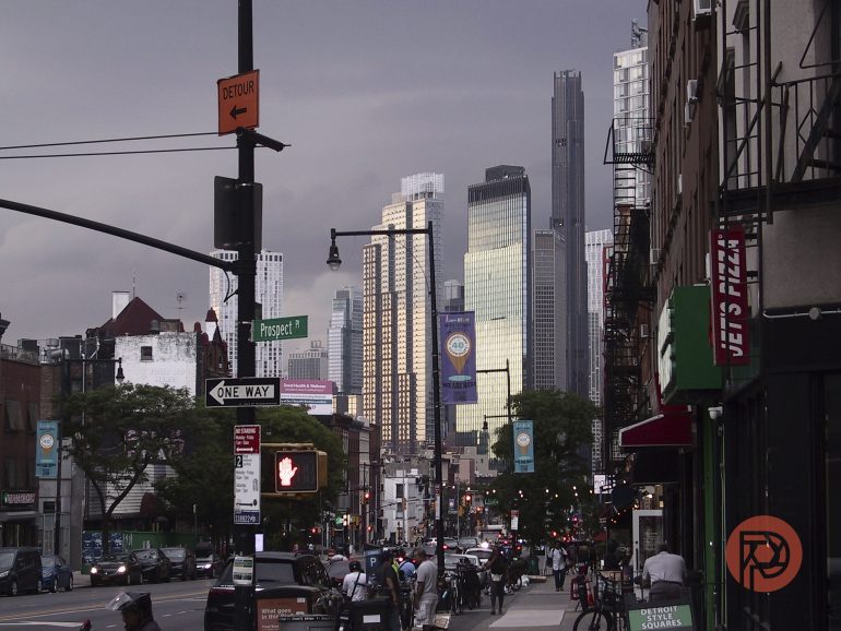 City street with cars, bikes, people, and tall buildings under a cloudy, darkening sky at sunset.