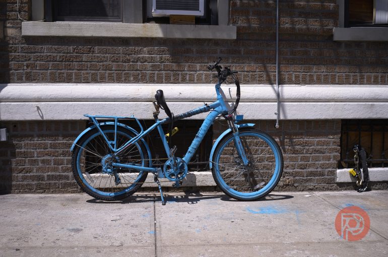 A blue-painted bicycle locked to a metal pipe on a sidewalk beside a brick building.