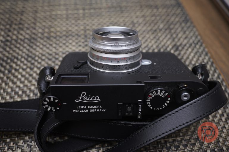 Leica camera with attached lens and strap resting on a textured woven surface.