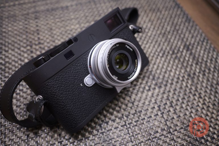 Black camera with a silver lens rests on a textured brown placemat, red logo in the bottom right corner.