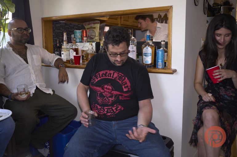 Three people sit by a bar with drinks, while another stands in the background. Bottles line the counter.