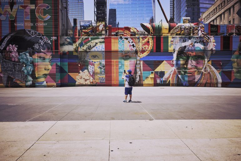 Child wearing a backpack stands alone, photographing a colorful mural with faces on a city wall.