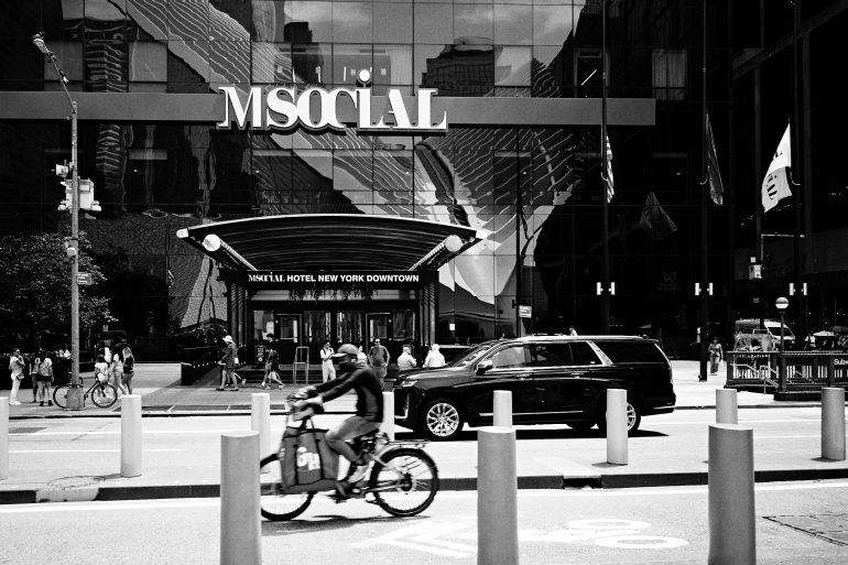 A person rides a bicycle past the MSocial Hotel in New York City; people and cars are in the background.