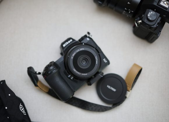 Viltrox 26mm F2.8 EVO Review: It Turned My Nikon Z7 II Into an EDC Camera A Nikon camera with lens and strap, lens cap removed, sits on a light surface beside another camera.