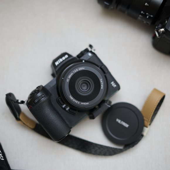 A Nikon camera with lens and strap, lens cap removed, sits on a light surface beside another camera.