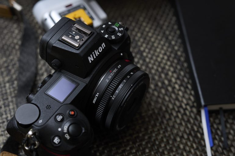A Nikon camera with a lens sits on a woven surface next to a notebook and camera battery.