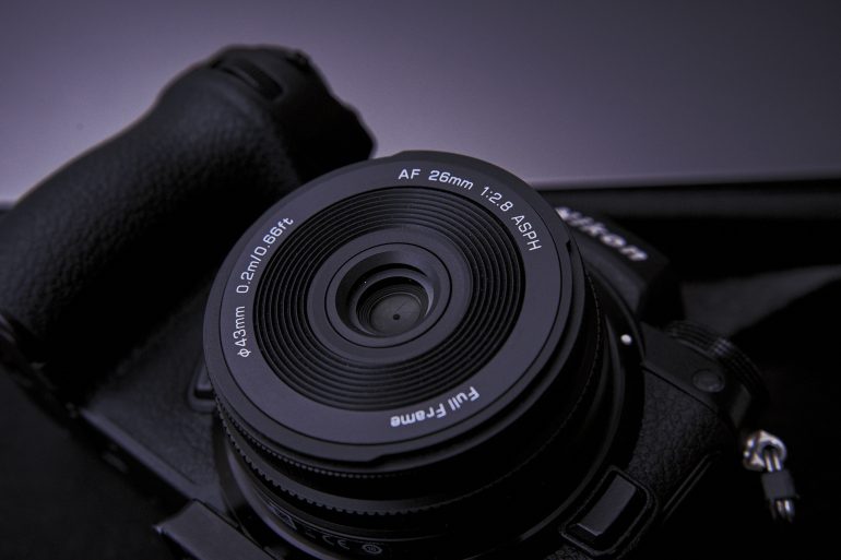 Close-up of a black digital camera with a 26mm f/2.8 ASPH lens against a dark background.