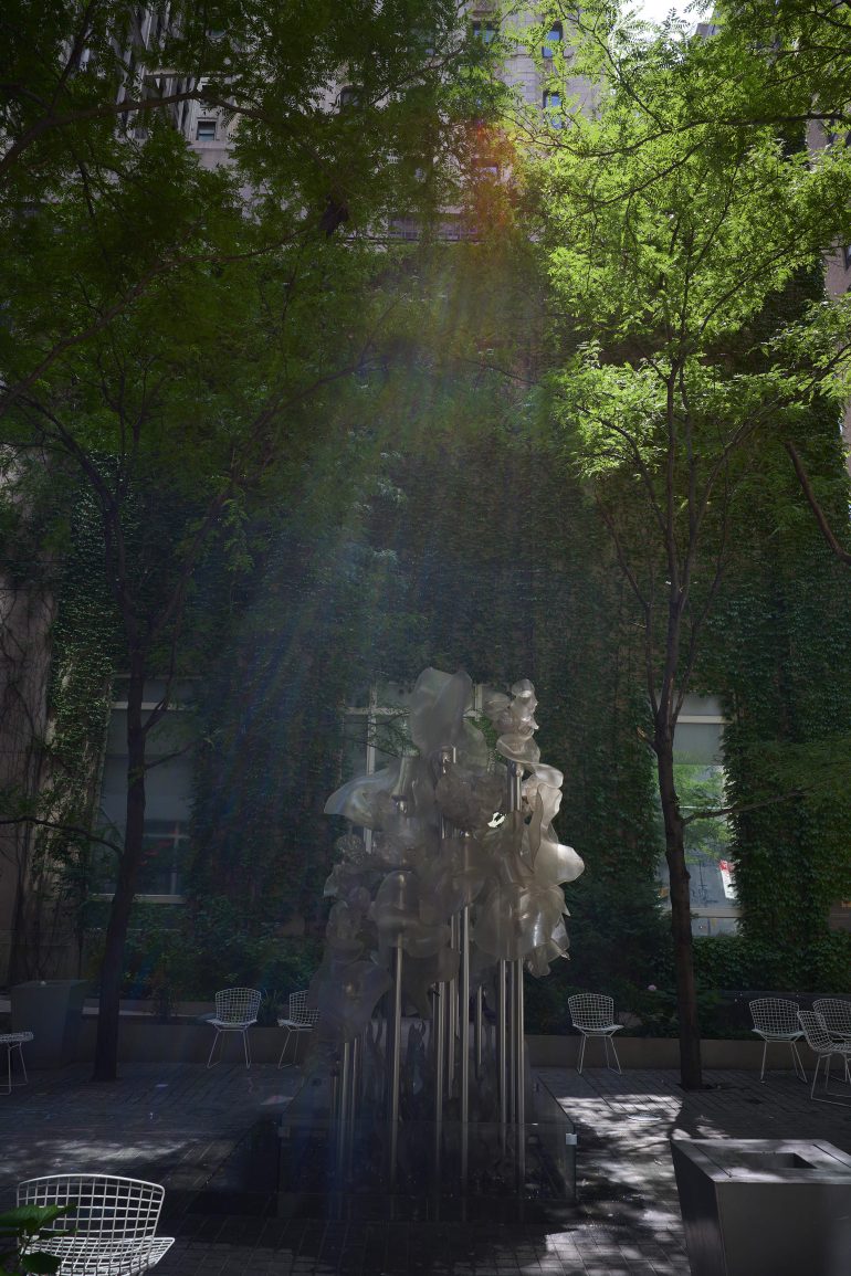 Sunlit courtyard with white sculptural fountain, metal chairs, and green trees against ivy-covered walls.