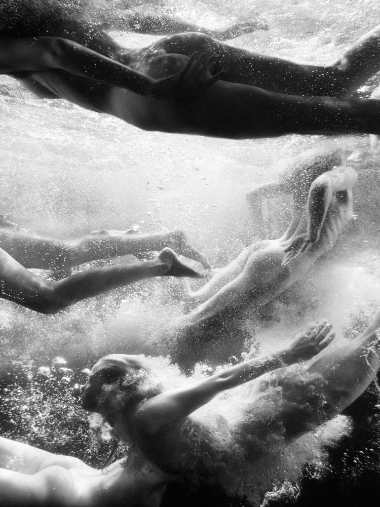 Four people swim underwater in black and white, surrounded by bubbles and light, captured in motion.