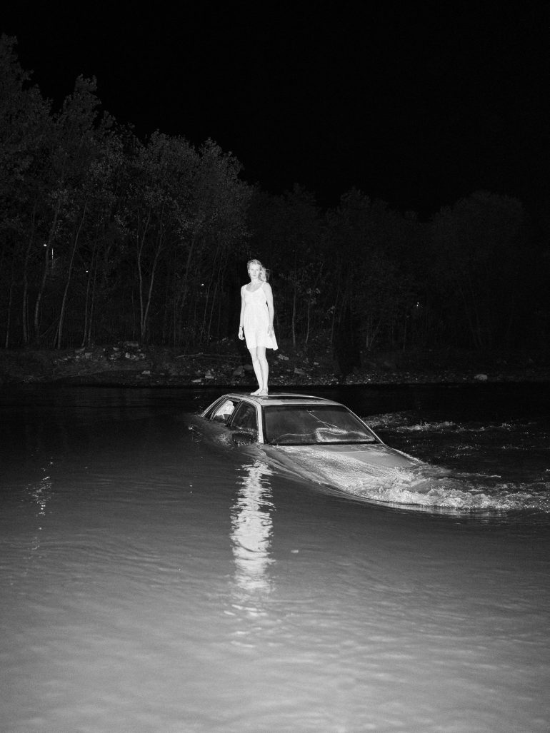 A woman in a dress stands on a partially submerged car in water at night, with trees in the background.