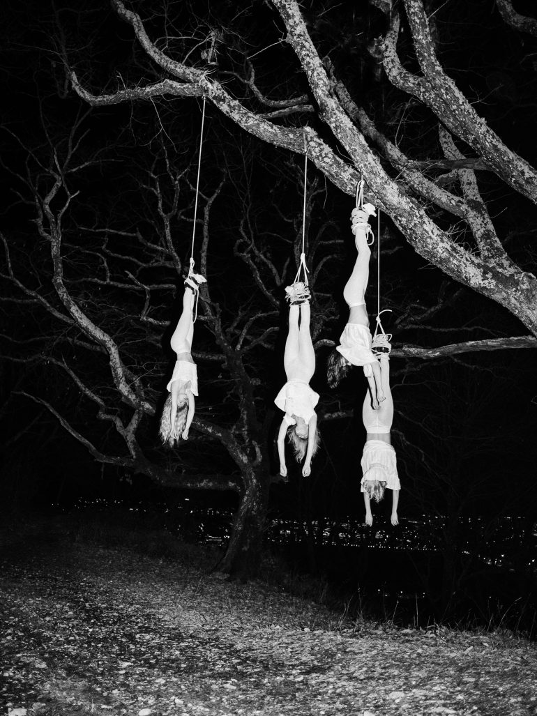 Three people hang upside down from tree branches at night, each tied at the ankles, city lights in the background.