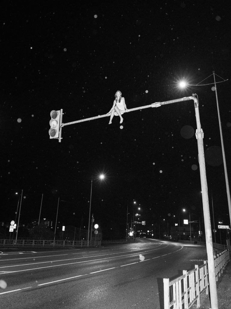 A person sits atop a traffic light pole above an empty road at night, with streetlights and rain visible.