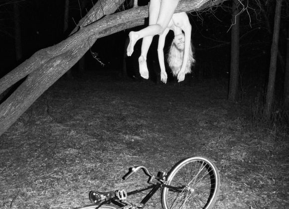 The Photographer That's Making You Look Twice at a Frame (NSFW) A person hangs nude from a tree branch at night, with a bicycle lying on the ground below.