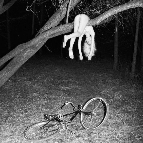 A person hangs nude from a tree branch at night, with a bicycle lying on the ground below.