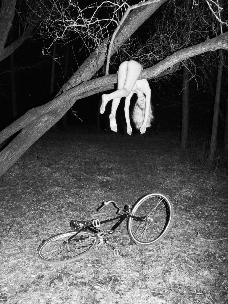 A person hangs nude from a tree branch at night, with a bicycle lying on the ground below.