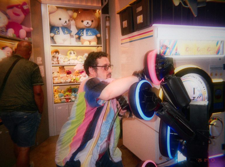 A man plays an arcade punching game, with plush toys displayed on shelves in the background.
