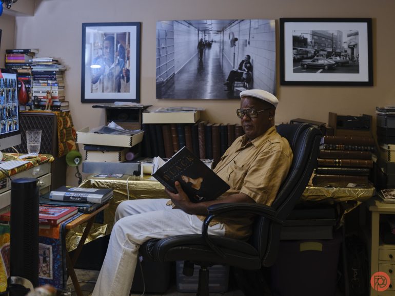 Man in beige clothes reads a book in a cluttered room with framed photos on the wall behind him.