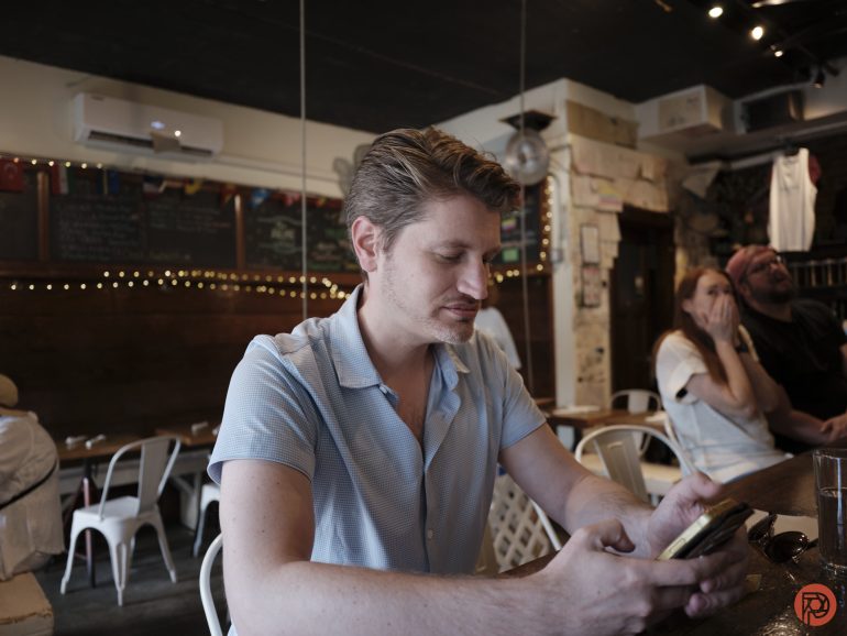 Man in a light blue shirt sits in a cozy café, looking at his phone; other patrons are in the background.