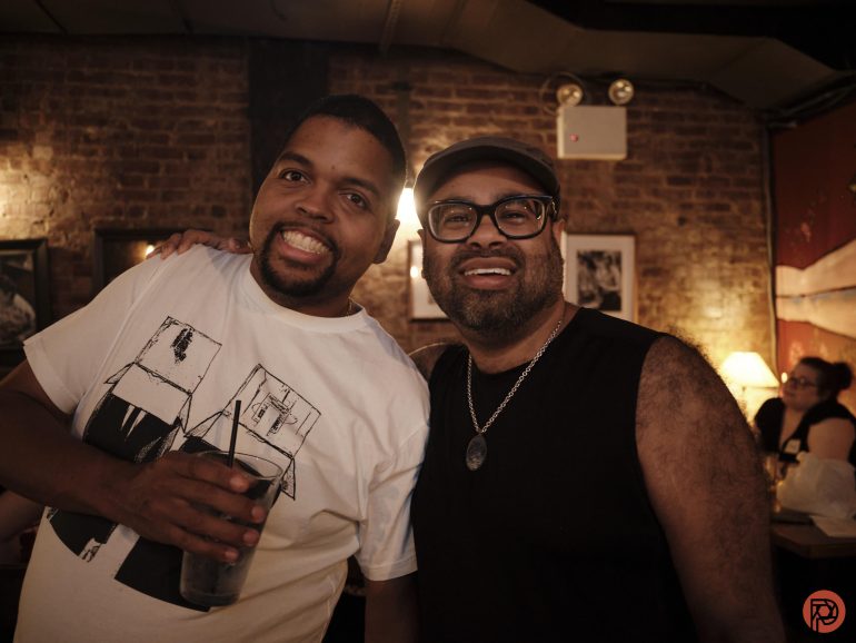 Two smiling men pose together in a cozy, dimly lit bar with brick walls and framed art.