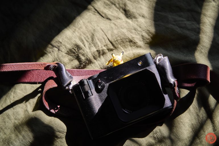A small Pikachu figure sits on top of a black camera with a brown strap on a green fabric background.