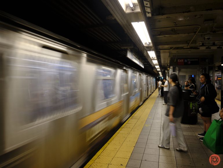 A subway train speeds by on a platform as people wait and stand under fluorescent lights.