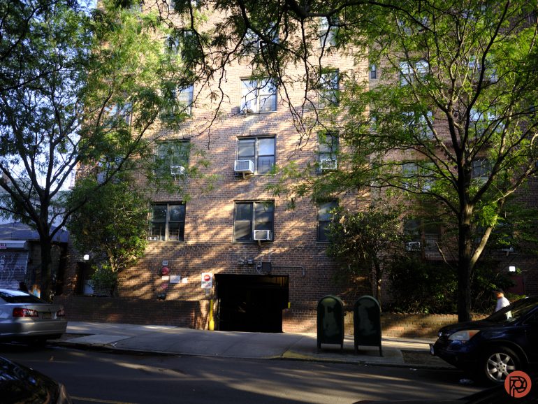 Brick apartment building with trees, parked cars, and a driveway entrance on a sunny day.
