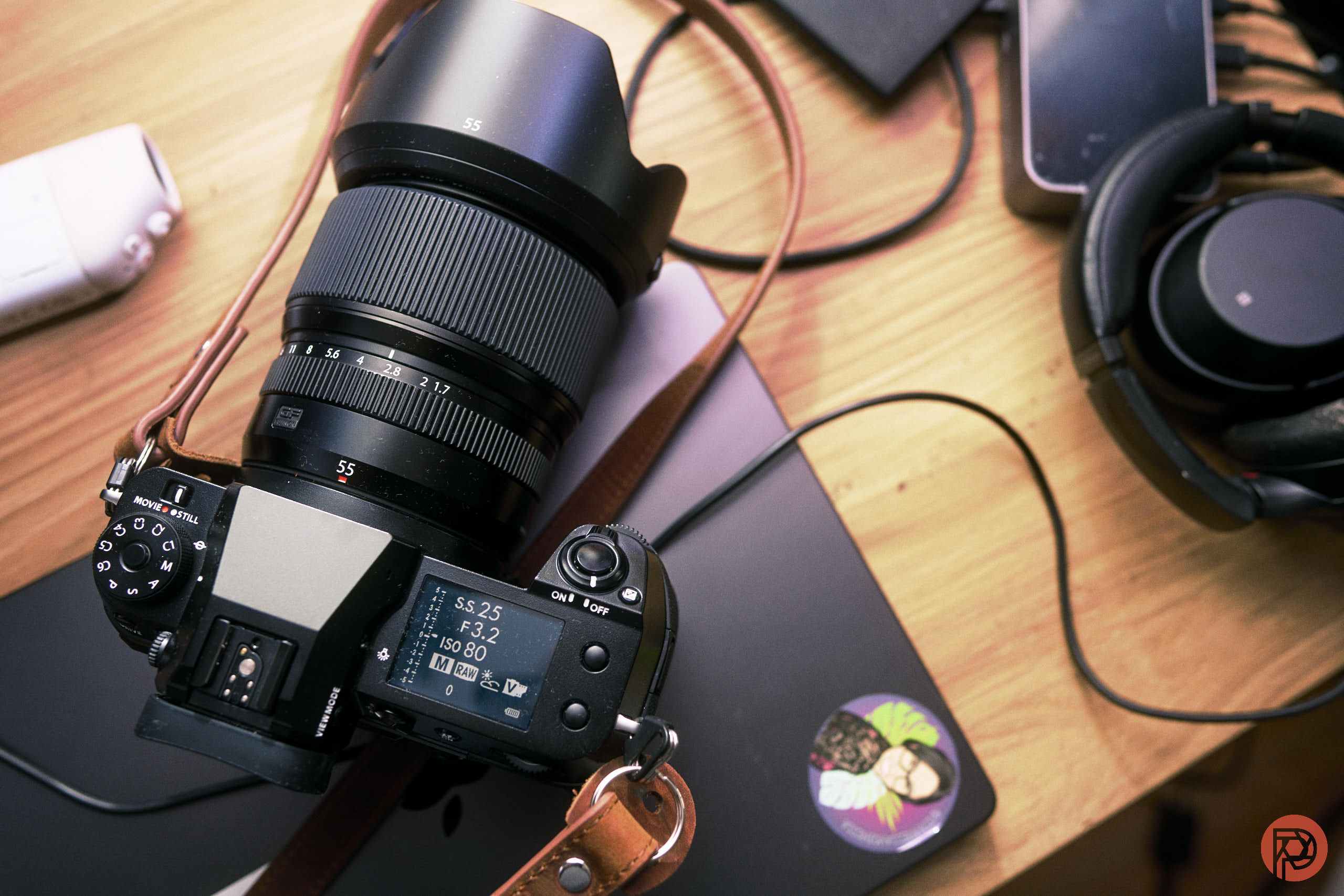 Should You Get the Fujifilm GFX 100S II in 2026? A digital camera on a desk with headphones, a phone, and a closed laptop nearby.