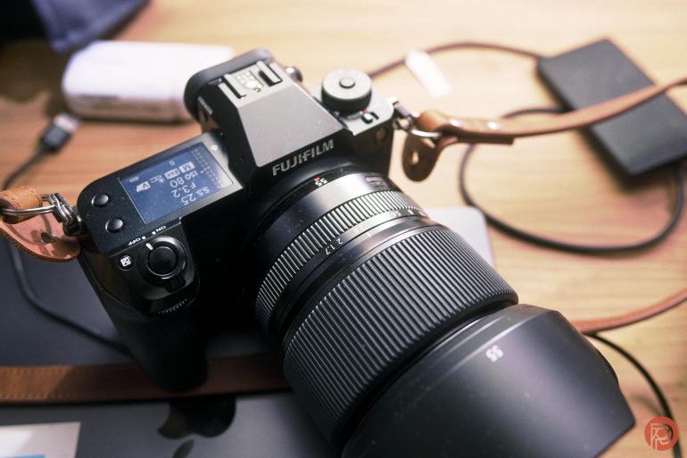 A black Fujifilm camera with a large lens sits on a wooden desk with accessories nearby.