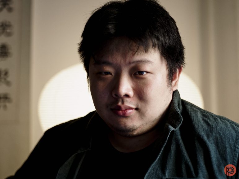 A person with short black hair and a green shirt looks at the camera, with a blurred light in the background.
