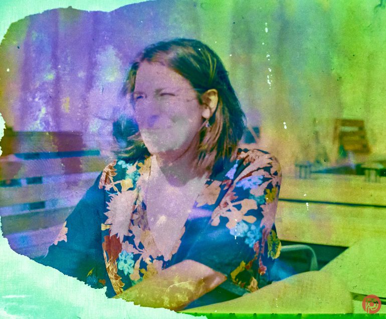 Woman in a floral dress sits at a desk, photo covered in bright paint splatters and blurs.