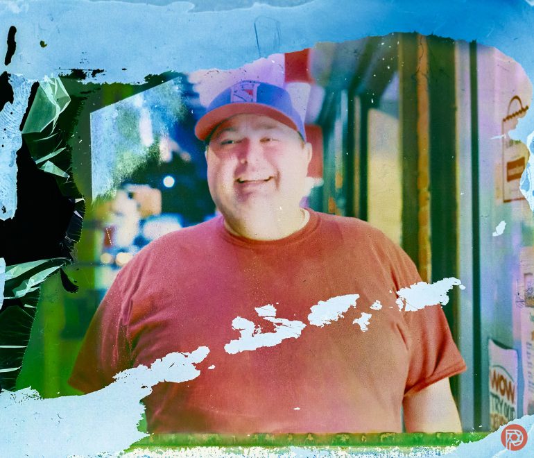 Smiling man in a red shirt and cap stands outside at night, with blue and green abstract paint effects.