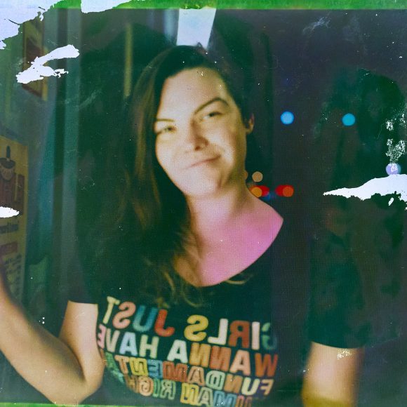 Smiling person with long hair wears a GIRLS JUST WANNA HAVE FUNDAMENTAL HUMAN RIGHTS shirt at night.