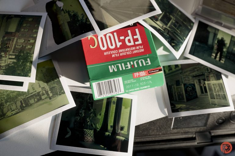 Scattered instant film photos and a Fujifilm FP-100C film box on a table, lit by sunlight.