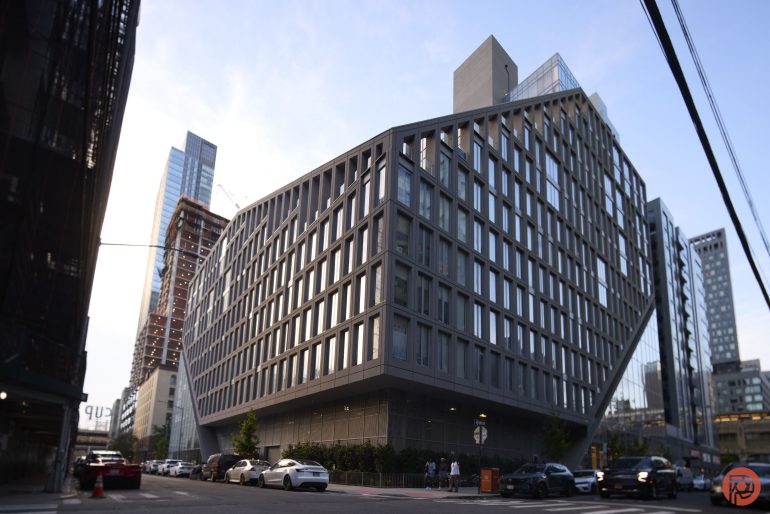 A modern, angular building with a grid-like facade stands on a city street lined with cars.