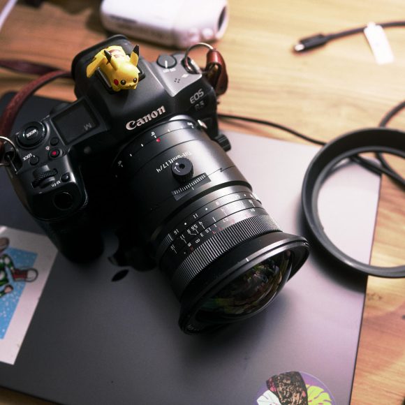 Canon camera with a Pikachu figure on top, resting on a closed laptop with stickers, on a wooden desk.