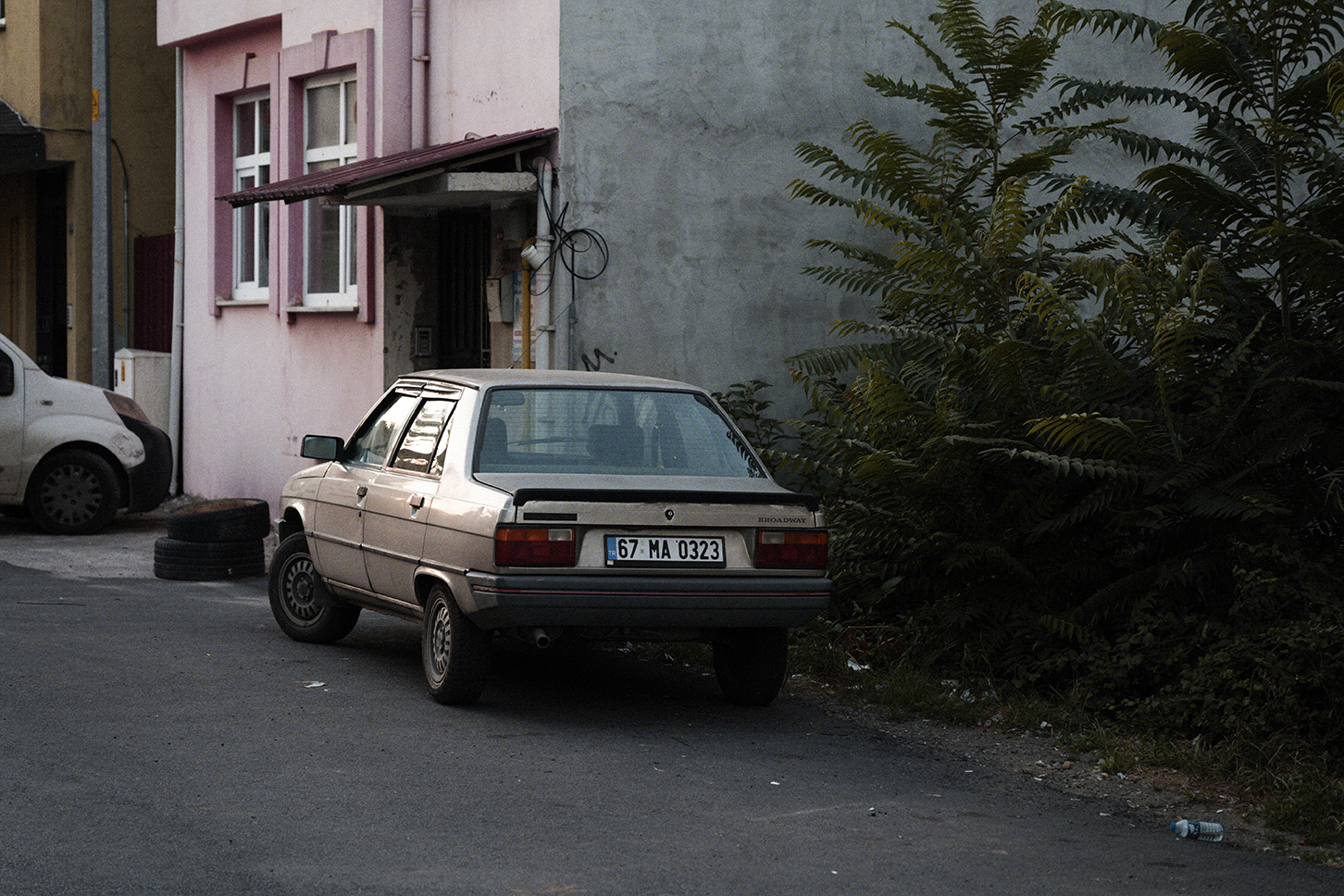 Old silver sedan parked near a pink building and large green bushes on a quiet street in the evening.