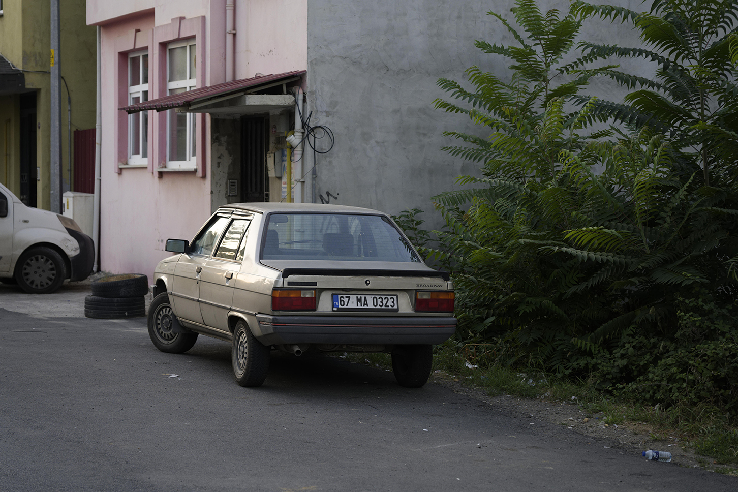 An old beige car is parked beside a pink building and green bushes on a quiet street.