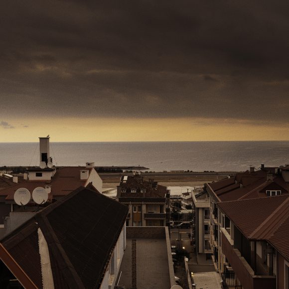 Red-roofed buildings overlook a calm sea under a dramatic, cloudy sunset sky.