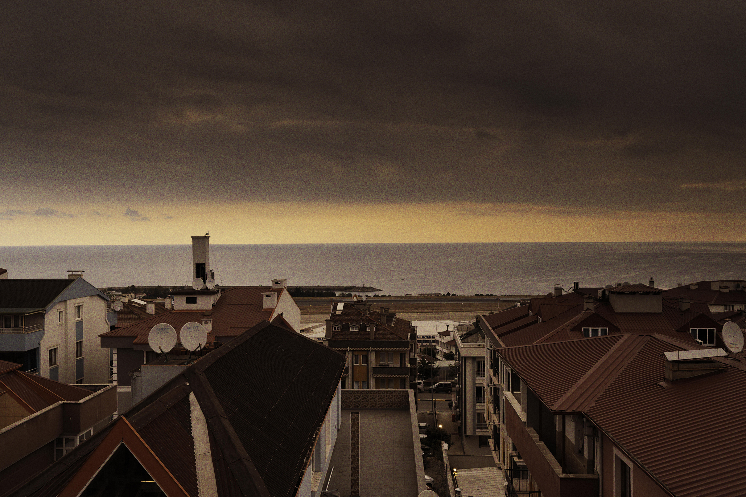 Red-roofed buildings overlook a calm sea under a dramatic, cloudy sunset sky.