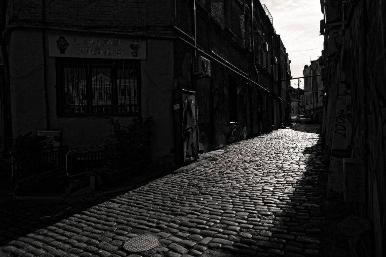 A cobblestone street in deep shadow, bordered by old buildings under a bright, overexposed sky.