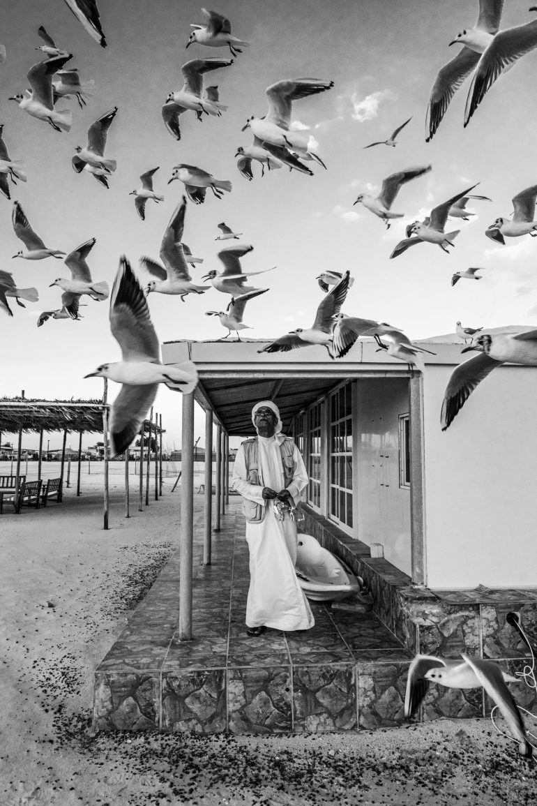A man in traditional attire stands on a porch as many seagulls fly overhead in black and white.