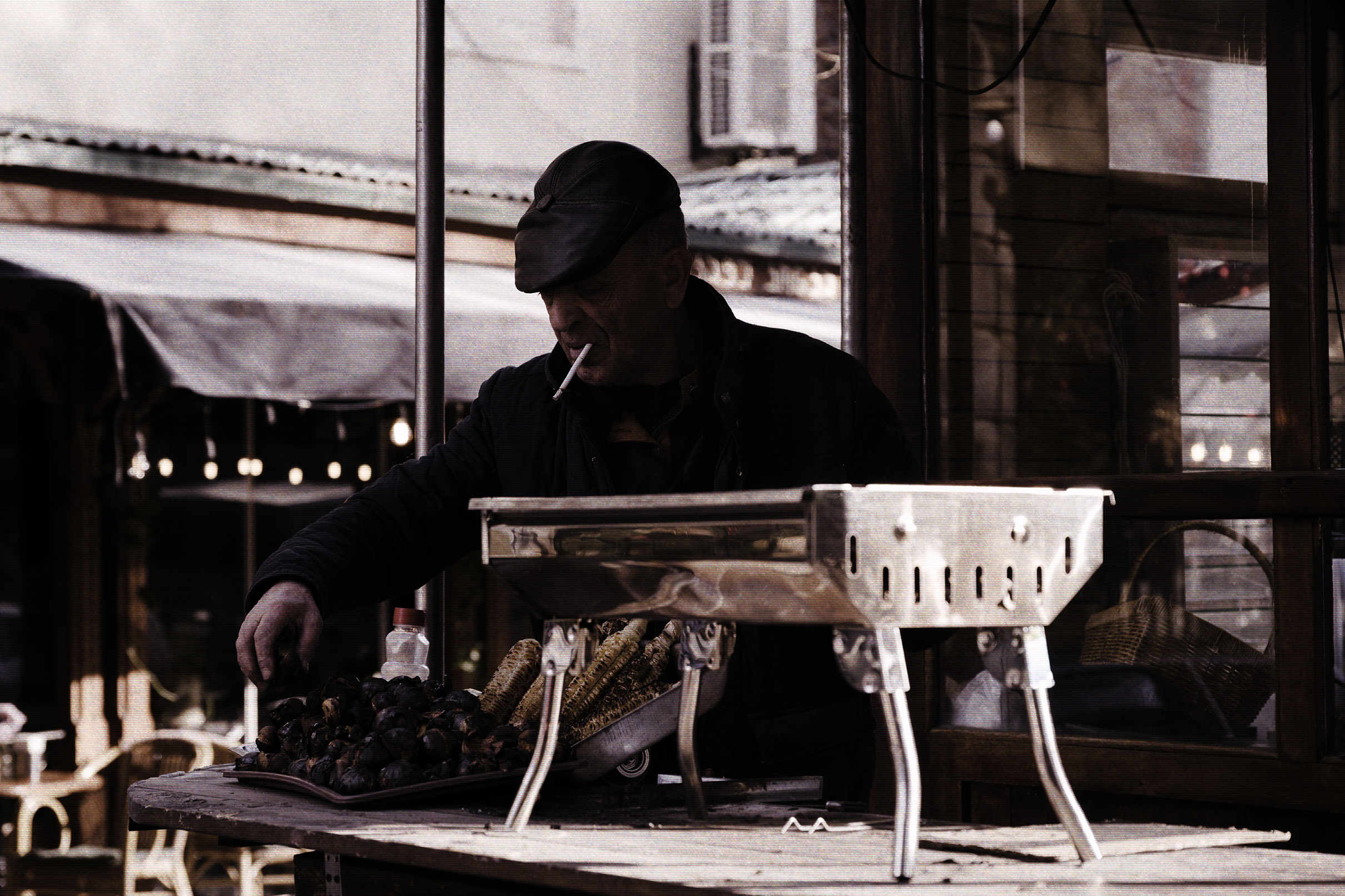 A man in a flat cap with a cigarette in his mouth cooks corn and chestnuts on a metal grill at an outdoor stall.