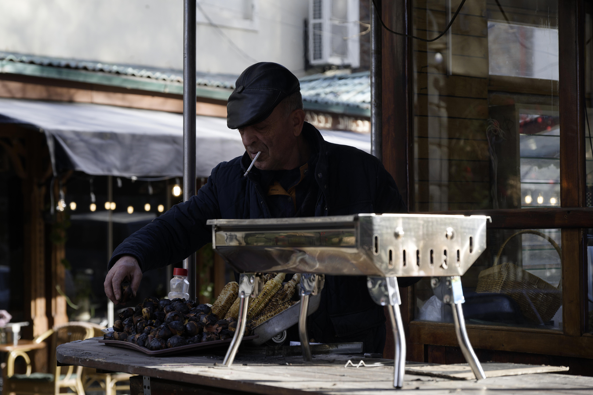 A man wearing a cap smokes while selling roasted chestnuts and corn from a street stall.