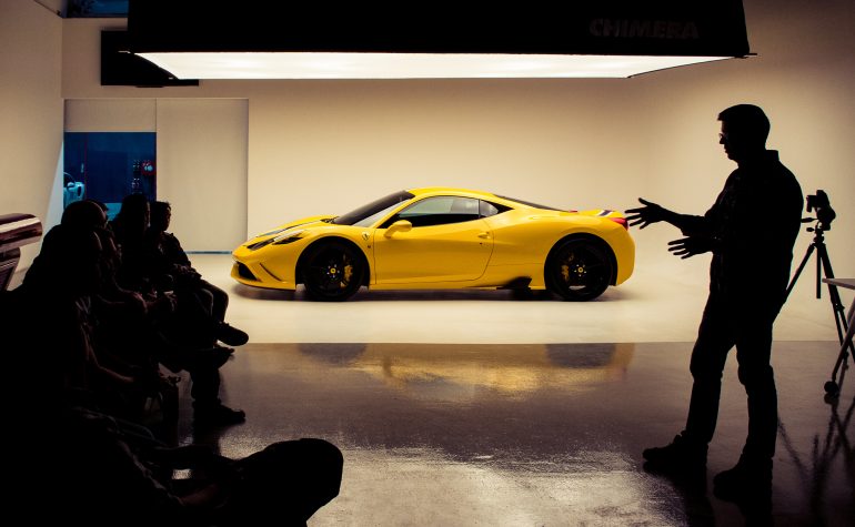 A yellow sports car in a studio, with a person speaking to a seated group in silhouette.