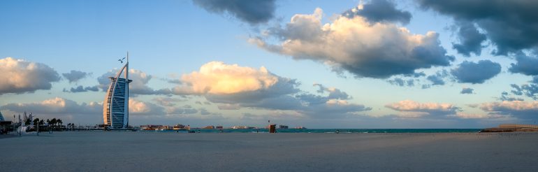Burj Al Arab hotel by the beach with scattered clouds in the sky and calm turquoise water.