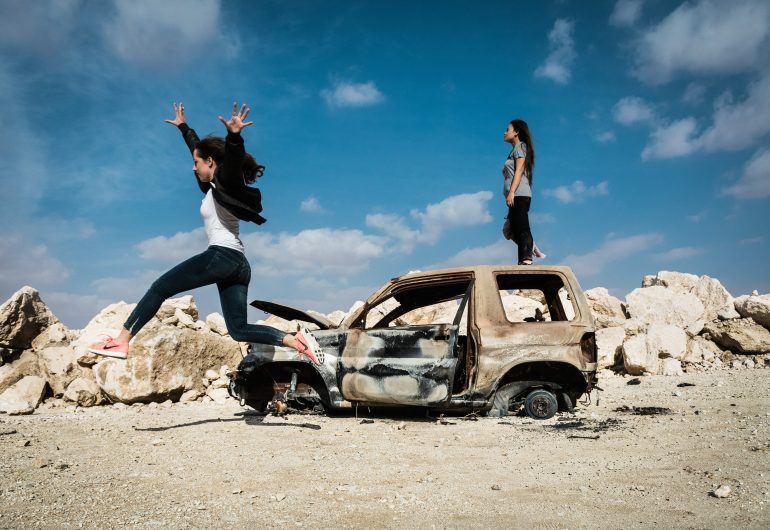 Two girls near a burnt-out car; one jumps mid-air, the other stands on top. Rocky landscape, blue sky.