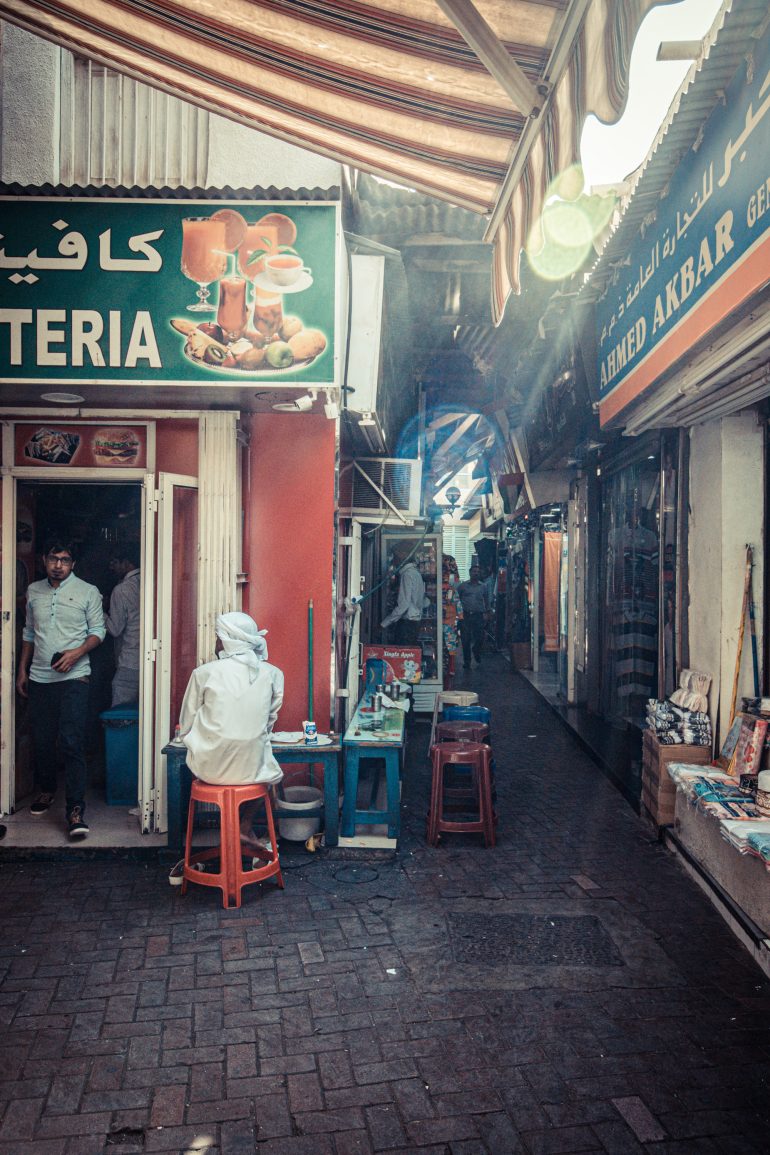 A narrow street market with shops, a seated man in white, and people walking through the alley.